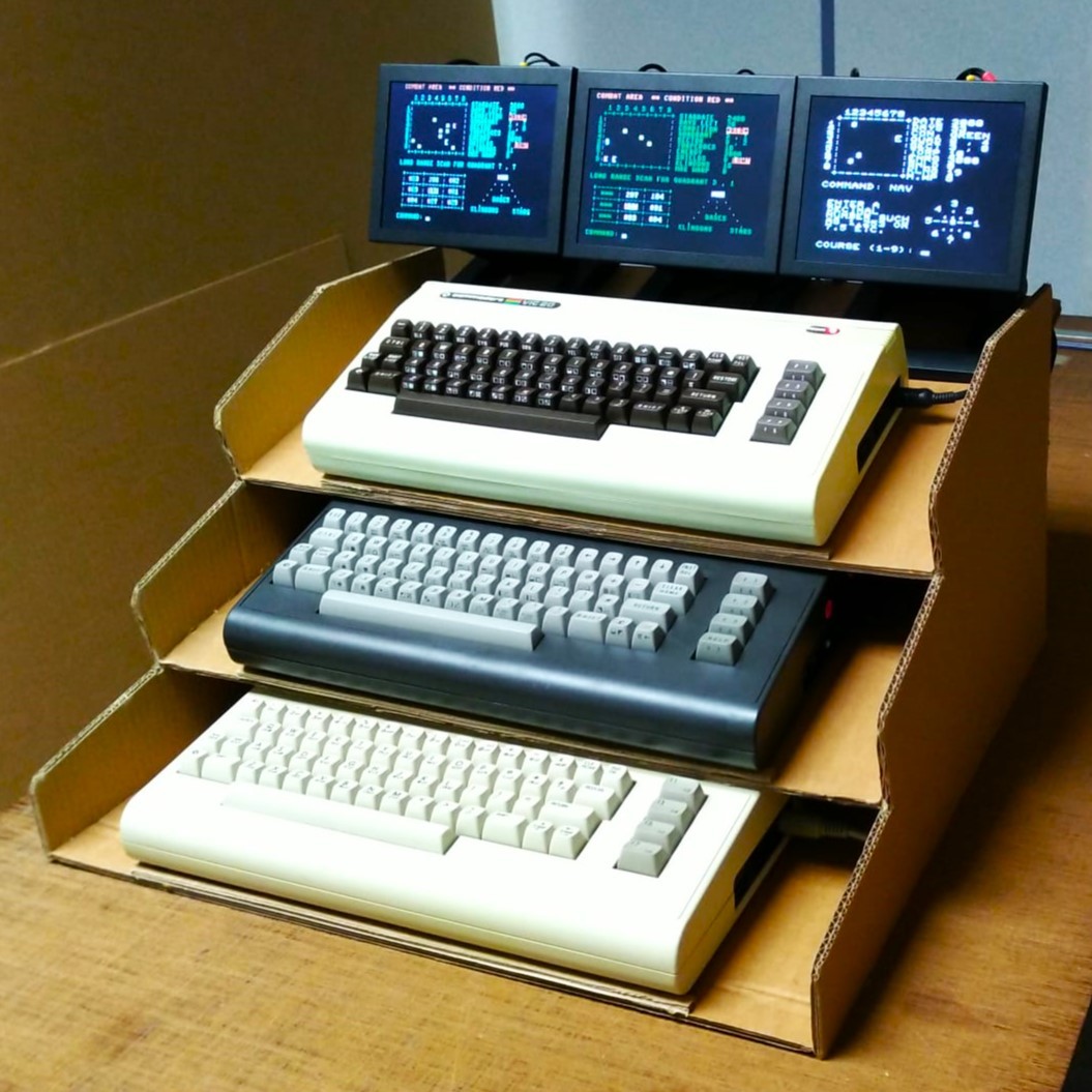 Vertically stacked and raked tier of 3 Commodore computers with 3 small LCD monitors on top. Prototype rack made of corrugated for proof of concept.