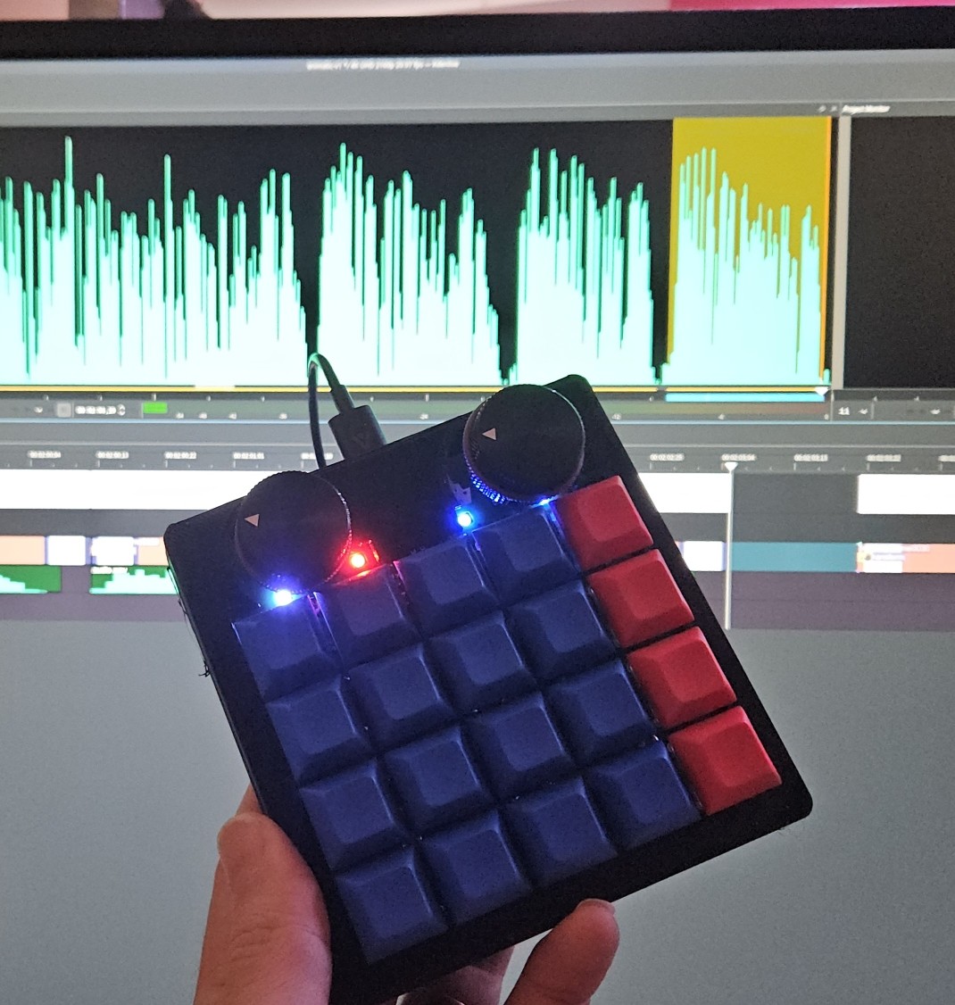 A 5x4 macropad with two large dials and some LEDs in front of a monitor with Kdenlive running.